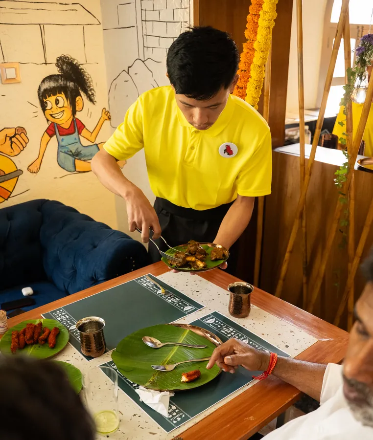 A server in a yellow Nati Republic tee setting a banana leaf at a table.