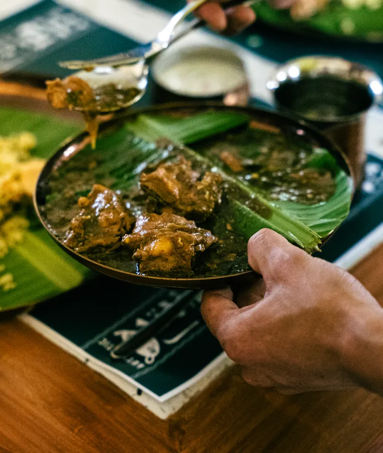 A bowl of mutton on banana leaf, a hand reaching in.